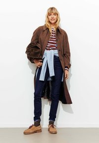 Manteau en laine brun à bouton devant, haut rayé rouge et blanc, pull bleu noué autour de la taille, jeans foncés et chaussures à lacets beiges.