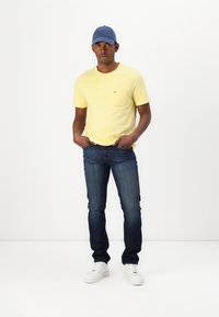 Gul bomulds T-shirt med brystlomme, parret med mørkeblå jeans og hvide sneakers. Bærer en blå kasket. Stående stilling.