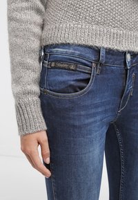 Närbild av en person som bär blå denimjeans och en grå stickad tröja, med vänsterhanden vilande vid sidan.
