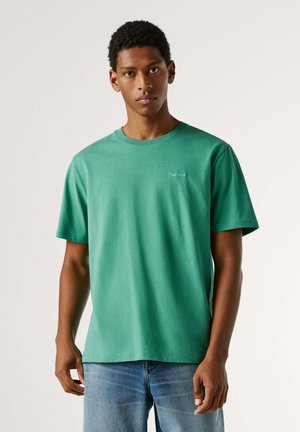 Jonge man die een groen T-shirt met korte mouwen en een blauwe spijkerbroek draagt, staand tegen een effen lichte achtergrond, met een neutrale uitdrukking frontal gericht.