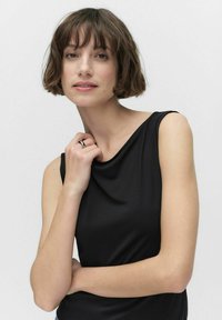 Svart ärmlös topp med draperad halsringning, mjuk tyg och åtsittande form. Modellen bär en silverring och har en kort bobfrisyr.