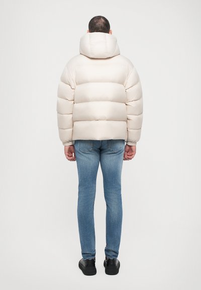 Polstret cremfaret pufferjakke med hætte, der har horisontale quiltede sektioner. Båret over blå jeans og sorte sko.