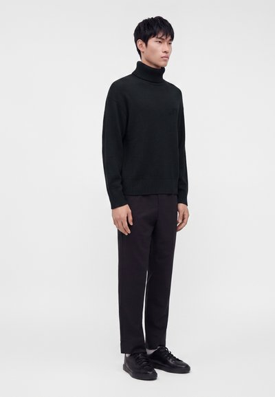 Jeune homme debout portant un pull à col roulé noir, un pantalon foncé et des baskets noires sur un fond clair uni.