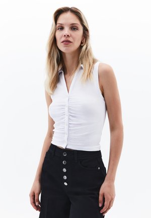 Weißes, geripptes, ärmelloses Shirt mit Kragen und Knöpfen an der Vorderseite, kombiniert mit hochtaillierten schwarzen Hosen mit silbernen Knopfakzenten.
