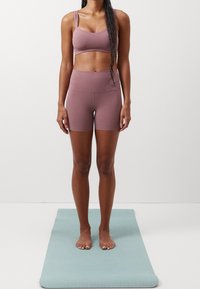 Vaaleanpunainen urheilurintaliivit ja shortsit, valmistettu pehmeästä, joustavasta materiaalista. Malli seisoo paljain jaloin vaaleansinisellä joogamatolla, neutraali tausta.