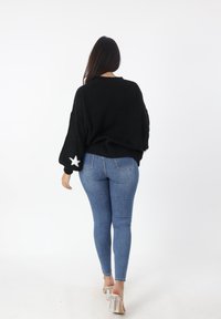 Zwarte oversized gebreide trui met brede mouwen, gecombineerd met blauwe skinny jeans. De trui heeft een witte steraccent op de manchetten.