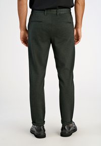 Tmavozelené elegantné nohavice s hladkou textúrou, úzkym strihom, dvoma zadnými vreckami a jemným prešívaním, skombinované s čiernymi topánkami.