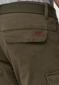 Nærbillede af olivenfarvede bukser med en klaplomme, der har et brunt "INDICODE EST. 1993" mærke samt et matchende vævet bælte.