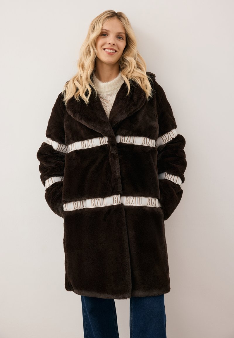 Bruine nepbontjas met grote kraag, witte merkbanden rond de taille en mouwen, en een relaxte pasvorm. Lange mouwen en zijzakken.