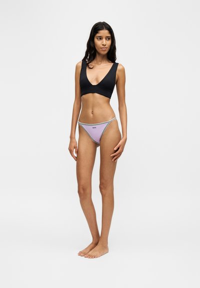 Femme debout pieds nus portant un crop top noir avec un décolleté en V profond et un bas de bikini lavande avec une ceinture blanche et grise sur fond blanc.