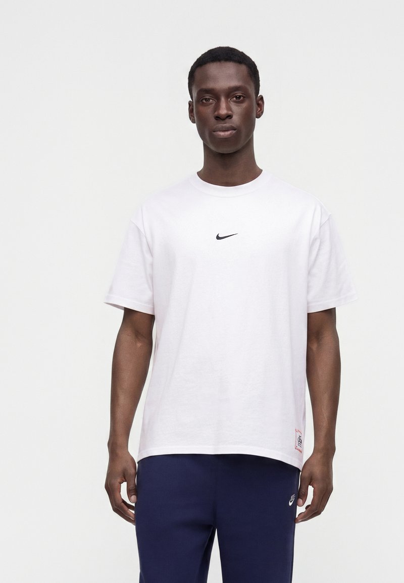 Ung mand iført en hvid Nike t-shirt og marineblå Nike bukser, står foran en ensfarvet lys baggrund.