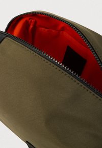 Τσάντα ελιάς με μαύρο φερμουάρ, που διαθέτει φωτεινή κόκκινη επένδυση στο εσωτερικό και ομαλή υφασμάτινη επιφάνεια. Ορατή οργανωτική τσέπη στο εσωτερικό.