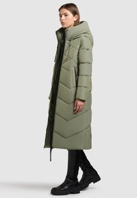 Ilgas pūkinis paltas alyvuogių žalia spalva su chrevrono siuvinėjimu, užtrauktukais uždaromomis kišenėmis ir aukštu apykakle. Dėvimas su prigludusiomis juodomis kelnėmis ir masyviu juodu avalyniu.