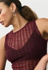 Burgundy top featuring a textured, pleated design with sheer sections. Model wears a decorative earring with pearl accents.