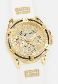 Orologio color oro con ghiera ricoperta di cristalli trasparenti, cinturino in silicone bianco, quadranti multifunzione e logo centrale "Guess".