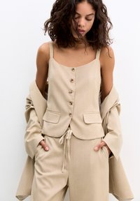 Ensemble en tissu beige côtelé comprenant un top sans manches avec des boutons sur le devant et deux poches, associé à un pantalon à cordon assorti et une veste.
