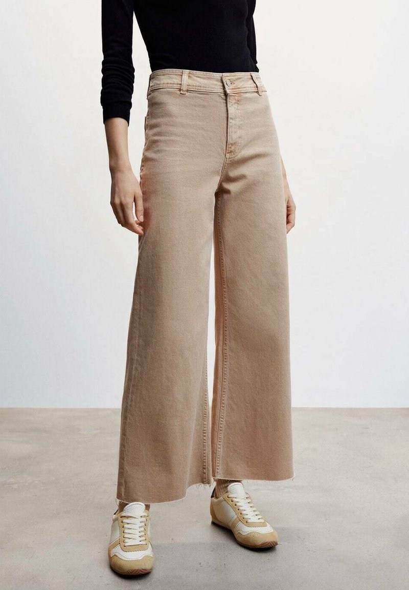 Personne portant un pantalon beige à jambes larges, des baskets blanches et beiges, et un haut noir à manches longues, debout sur un sol gris contre un mur blanc.