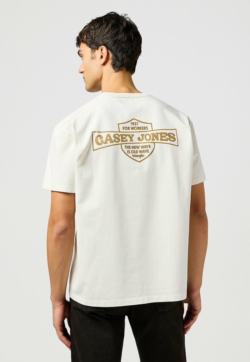 Camiseta de algodón blanca con un estampado gráfico marrón en la parte posterior que incluye el texto "CASEY JONES", un logotipo y un cuello redondeado.