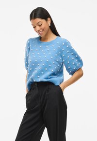 Suéter de punto azul claro con mangas cortas abullonadas, que presenta puntos texturizados blancos. Combinado con pantalones negros de cintura alta.