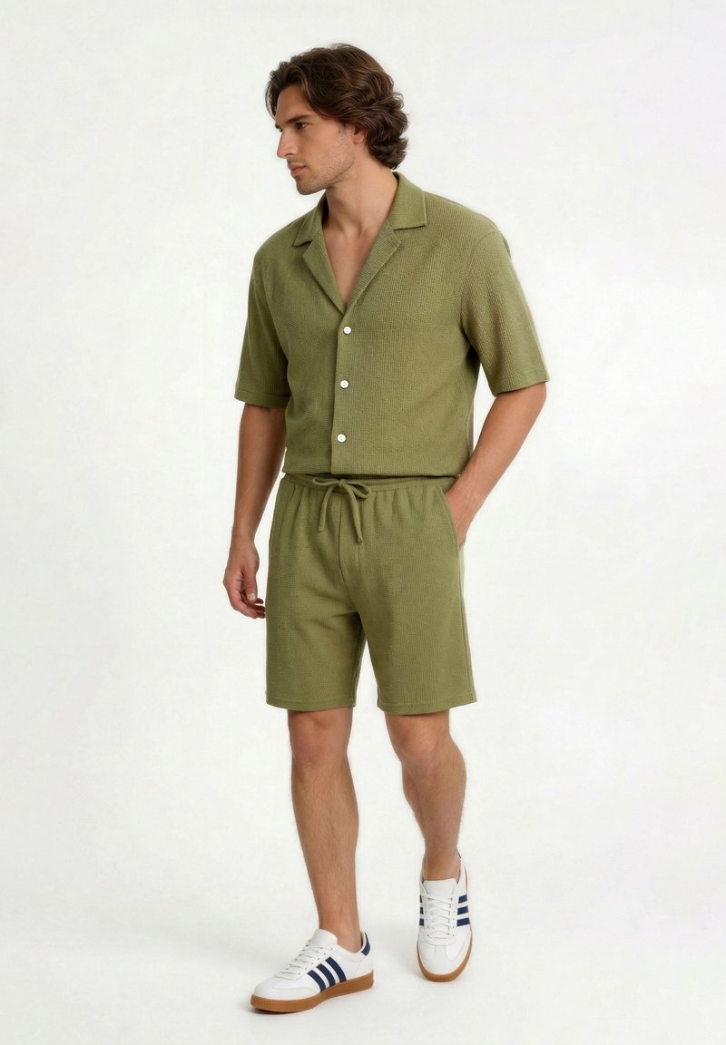 Mand iført olivengrøn kortærmet skjorte med knapper og matchende shorts med hvide sneakers går mod en ensfarvet baggrund.