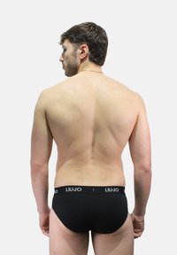 Mutandine nere con fascia in vita morbida che mostra il nome del marchio "LIU JO". Il modello è ritratto di spalle, con la schiena nuda rivolta verso la fotocamera.