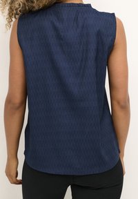 Marinblå ärmlös topp med texturerat mönster, rundad fåll och volangdetalj vid halsen. Tillverkad av lättviktig tyg.