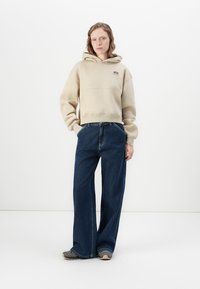 Felpa beige con cappuccio, dotata di tasca frontale e logo, abbinata a jeans a gamba larga blu scuro e sneakers grigie su uno sfondo bianco uniforme.