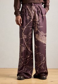 Pantalones de pierna ancha en tela de color marrón oscuro con un estampado de mapa en dorado que contrasta. Textura suave con detalles plisados y bolsillos laterales.