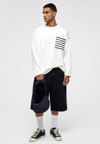 Witte lange mouwshirt met een zwart gestreepte zak, gecombineerd met zwarte denim shorts, witte sokken en zwarte sneakers, met een grijze tas in de hand.
