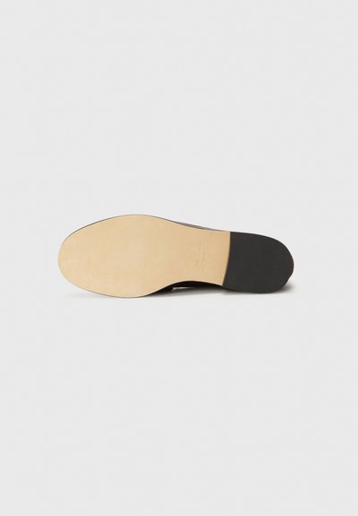 Zapato de cuero negro con una textura suave, que presenta una suela de goma plana de color claro. La suela tiene una punta redondeada y un detalle mínimo.
