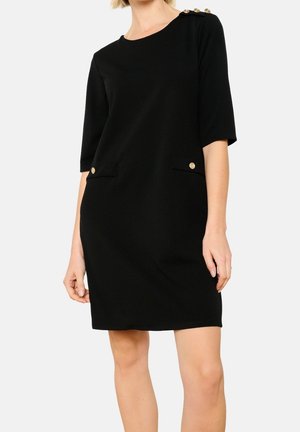 Zwarte jurk tot op de knie met halflange mouwen, gouden knopen op de schouder en voorzakken, gedragen door een staand persoon.