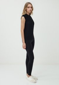 Una giovane donna si trova di profilo, indossando un top nero aderente e leggings, abbinati a sneakers bianche, su uno sfondo bianco uniforme.