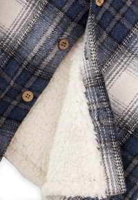 Kockovaná deka s modrými, šedými a bielymi vzormi. Jedna strana má jemný, biely, fleece-štýl textúry; obrácená strana má flanelovú úpravu s akcentmi gombíkov.