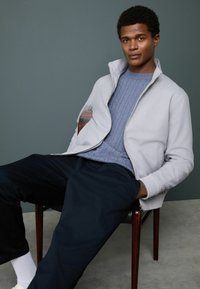 Seated man wearing a light gray zip-up jacket, blue cable-knit sweater, dark pants, and white socks against a gray wall and concrete floor.