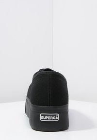 Baskets en toile noire avec une surface texturée, bout arrondi et semelle en caoutchouc. L'arrière présente une plaque logo argentée "SUPERGA".