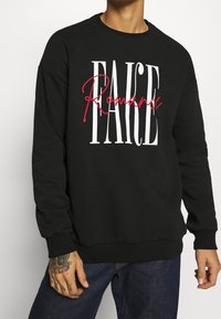 Persona vistiendo un sudadera negra con el texto blanco "FAKE" y rojo "Romance", combinada con jeans azul oscuro y un tatuaje en la mano visible en la mano izquierda.