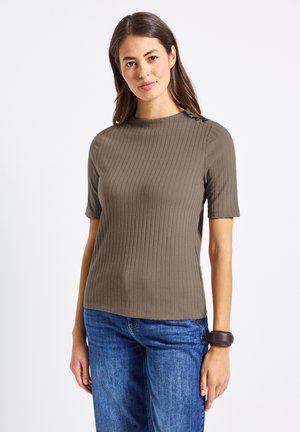 Žena v ribbed, krátkém rukávu taupe topu s knoflíky na ramenech a modrých džínách, stojící na jednoduchém bílém pozadí.