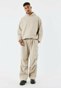 Felpa oversize beige con cappuccio e logo frontale, abbinata a pantaloni cargo coordinati con ampie tasche e vestibilità comoda. Scarpe nere.