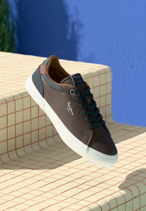 Brauner Ledersneaker mit weißer Sohle und schwarzen Schnürsenkeln, der auf cremefarbenen Treppen mit roten Gittern vor einer blauen Wand liegt.