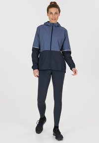 Giacca a vento leggera bicolore con cappuccio; parte superiore in azzurro chiaro, parte inferiore in blu navy scuro; dotata di zip e dettagli riflettenti.