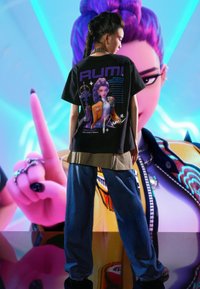 Fekete grafikai mintás póló "RUMI" felirattal és karakter dizájnnal a hátán, széles fazonú kék farmerral párosítva, alatta kockás ingréteggel.