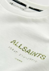 Valkoinen puuvillapaita, jossa on vihreää tekstiä "ALLSAINTS" ja "LONDON" teksturoidulla kuviolla. Pyöreä kaula-aukko, sileä pinta.