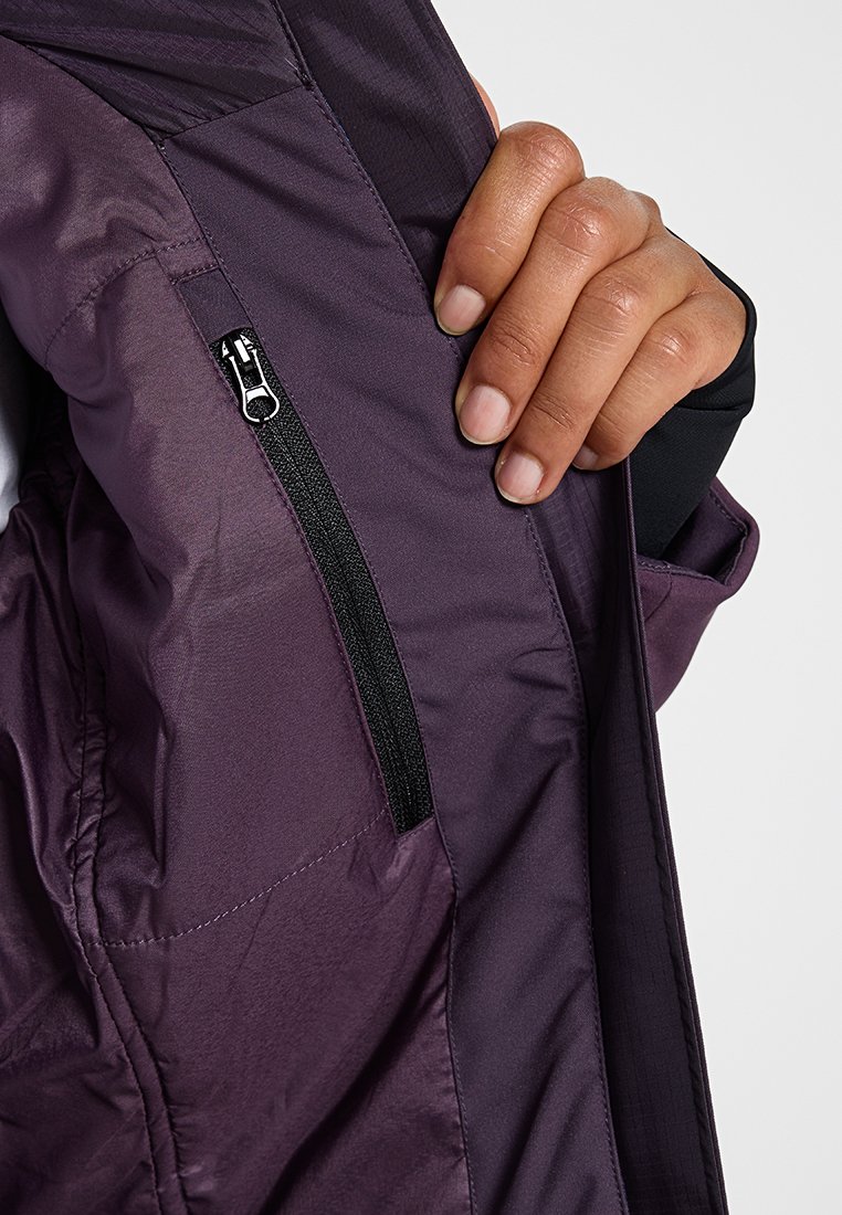 Mano che apre la tasca di un giubbotto viola mostrando una zip verticale nera con un tirante argentato su sfondo bianco.