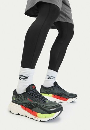 Piernas con leggings negros, calcetines blancos de Reebok y zapatillas deportivas negras de Reebok con suela multicolor (amarillo, rojo, blanco).