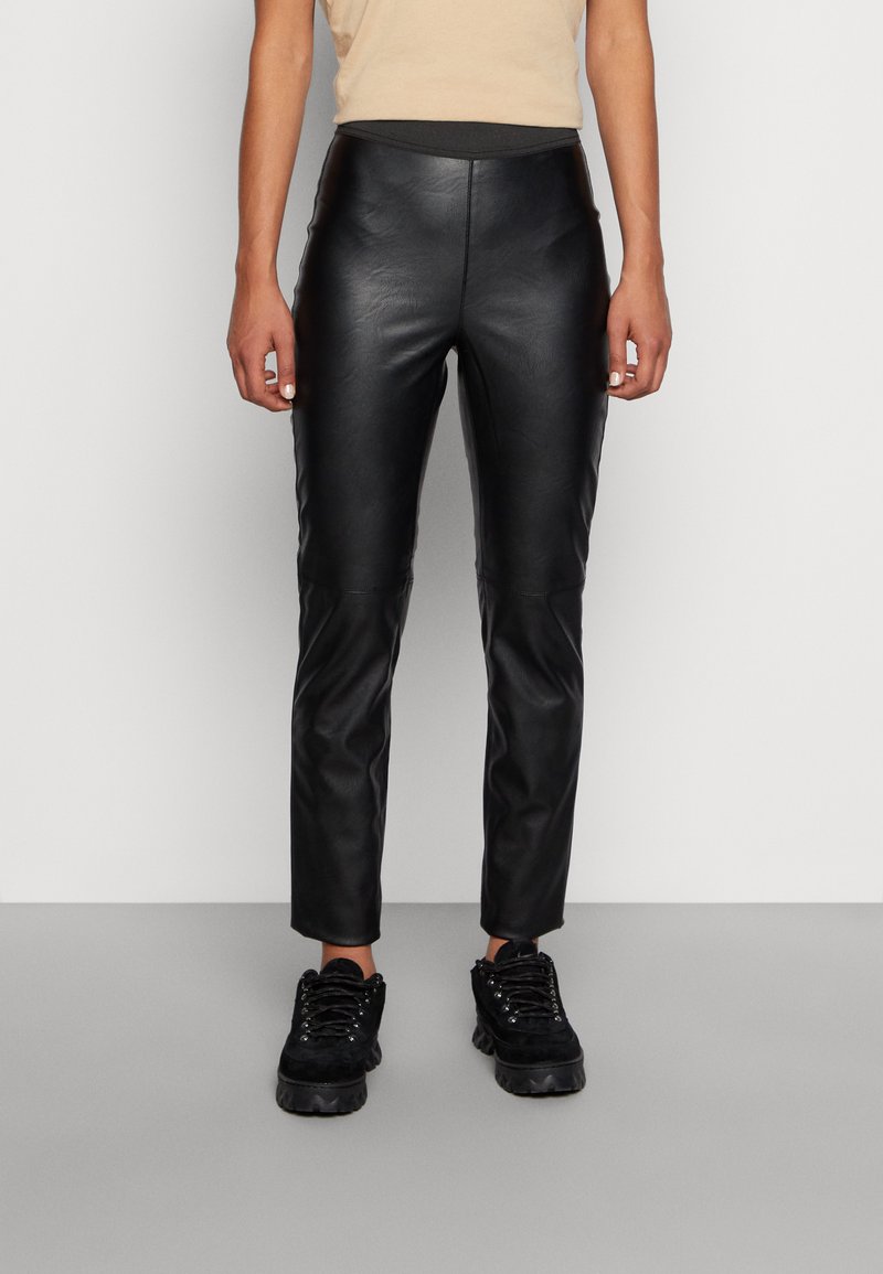 Calças de couro preto com um corte justo, apresentando uma textura suave, cintura alta e detalhes costurados, combinadas com sapatos chunky pretos.