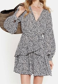 Svart klänning med vitt blommönster, långa vida ärmar, v-ringning och lager av volanger vid midjan. Kombinerad med en randig tygpåse.