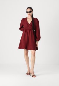 Rochie cărămizie cu decolteu în V și talie strânsă, având mâneci lungi tip puff. Modelul poartă ochelari de soare negri și sandale cu toc cu model.