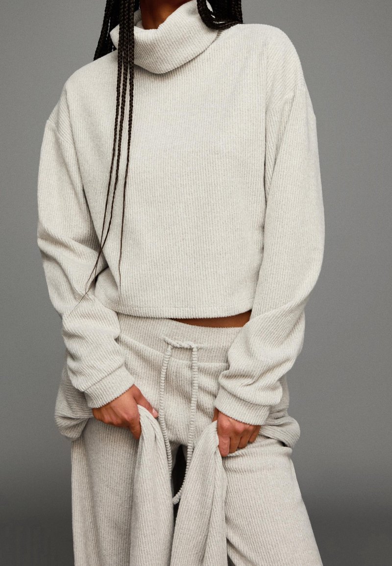 Personne portant un pull à col roulé côtelé beige clair et un pantalon à cordon assorti, tenant un tissu au niveau des cuisses sur un fond gris.
