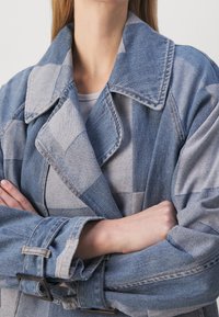 Denim jacket featuring a patchwork design in varying shades of blue, with a wide collar, belted waist, and textured fabric.