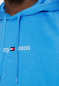 Gros plan sur un sweat à capuche bleu avec cordons et texte brodé « my » et « jeans » séparés par un logo rectangulaire rouge, blanc et noir.